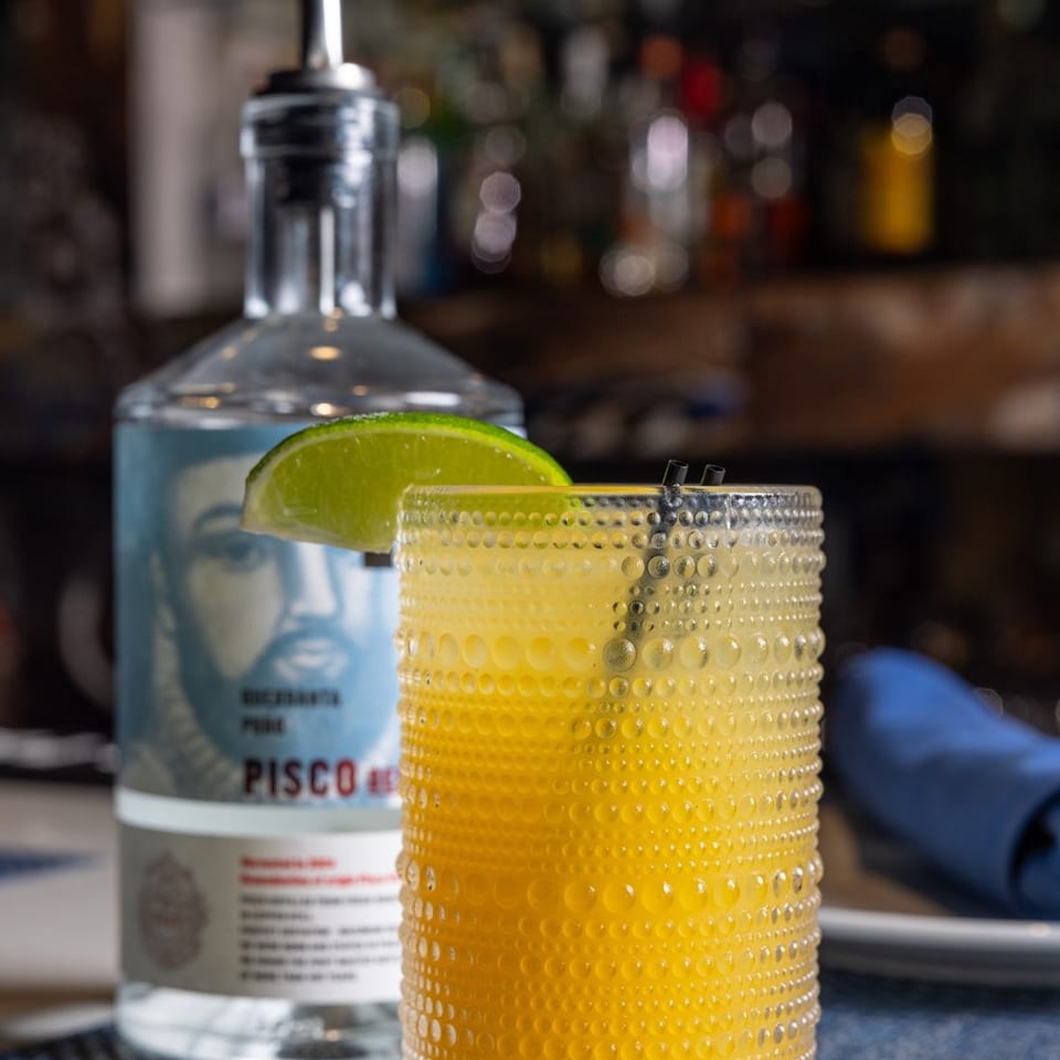 Textured glass of golden citrus cocktail garnished with lime, beside a bottle of pisco on a dimly lit bar.