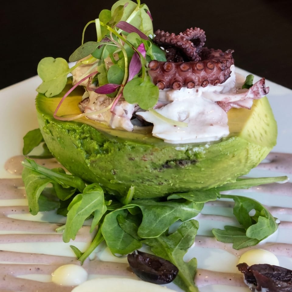 Stuffed avocado half topped with grilled octopus, microgreens, and vegetables on a white plate drizzled with sauce.