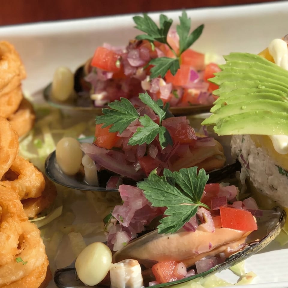 Colorful seafood platter featuring fried rings, mussels with salsa, avocado slices, and a layered yellow potato dish garnished with herbs.