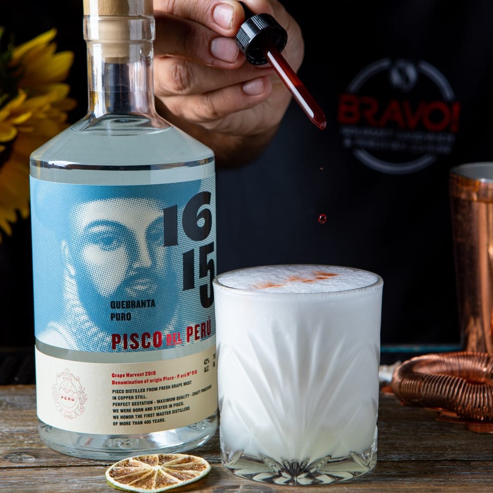 Bartender holds a dropper over a frothy pisco cocktail beside a bottle of Peruvian pisco, dried lime, and copper barware.