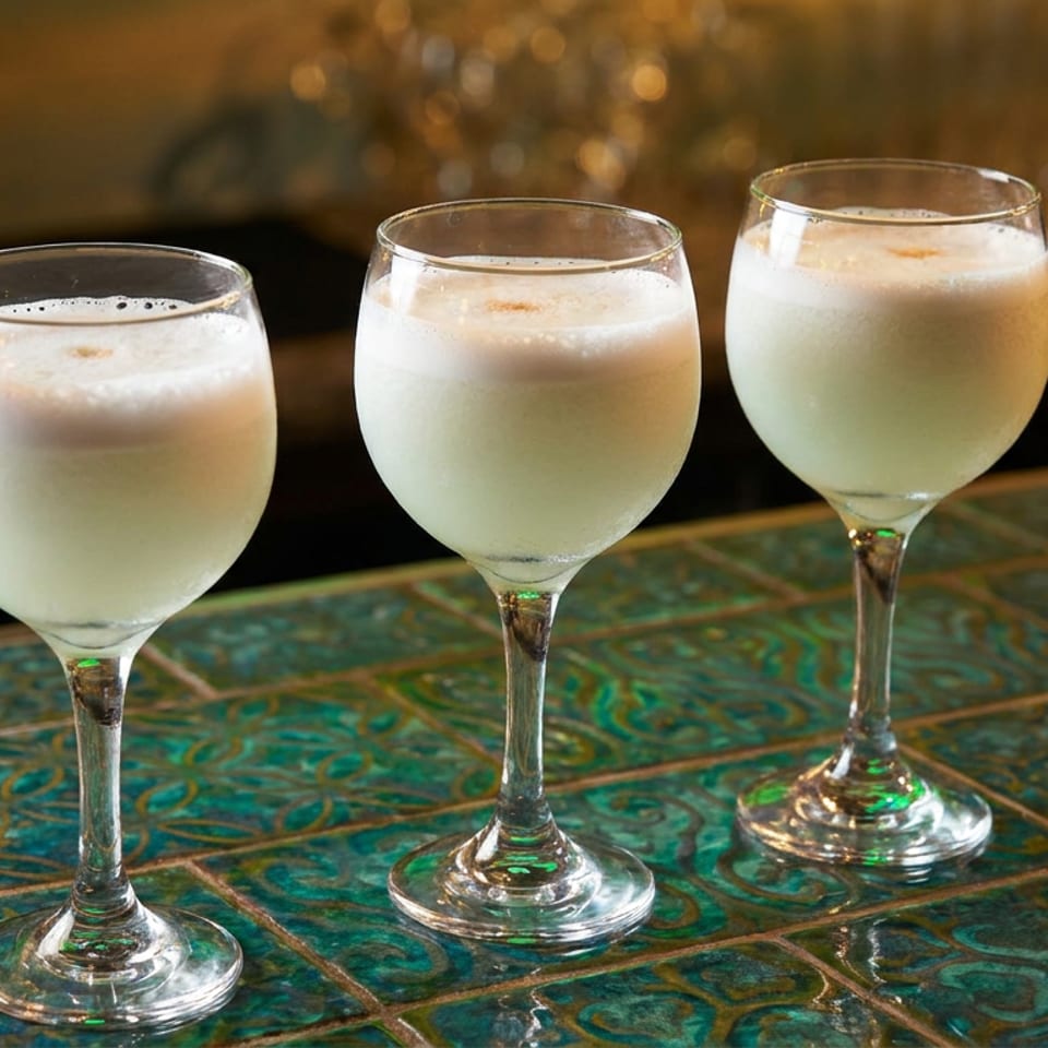 Three stemmed glasses filled with frothy white cocktails sit on a colorful patterned tile bar in a warmly lit restaurant.