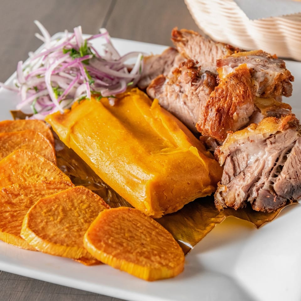 Plate of roasted pork, golden plantains, yuca, and shredded purple cabbage salad.