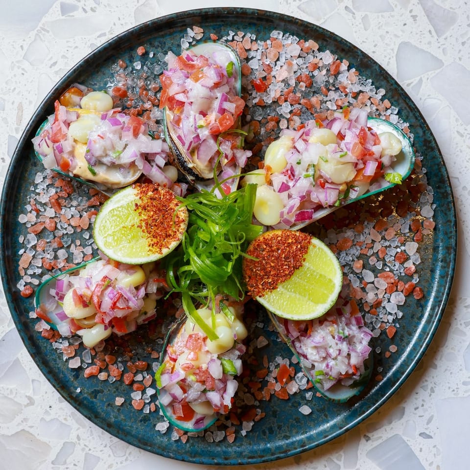 Five mussel shells topped with diced red onion and tomato, served on crushed ice with lime wedges and seaweed garnish.