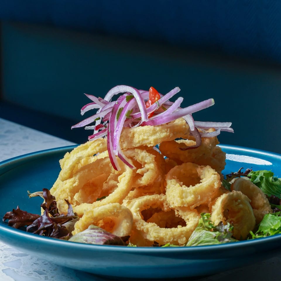 Crispy fried calamari rings topped with sliced red onions and tomatoes, served with mixed greens on a blue plate.